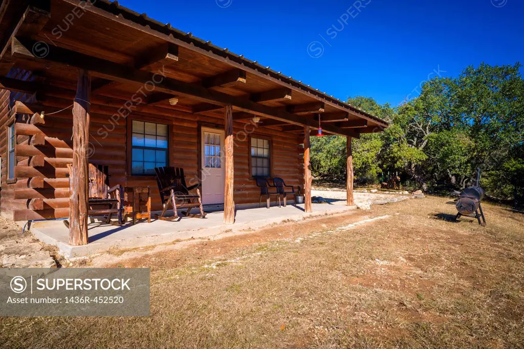 Exterior of rustic farm log house with wooden rocking chairs on the fron deck. Barbecue smoker in the front yard. Texas, USA.