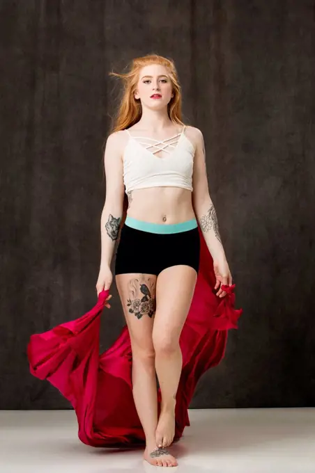 Young female dancer with long red hair and tattoos, holding red fabric that trails behind her as she walks forward on a white floor.