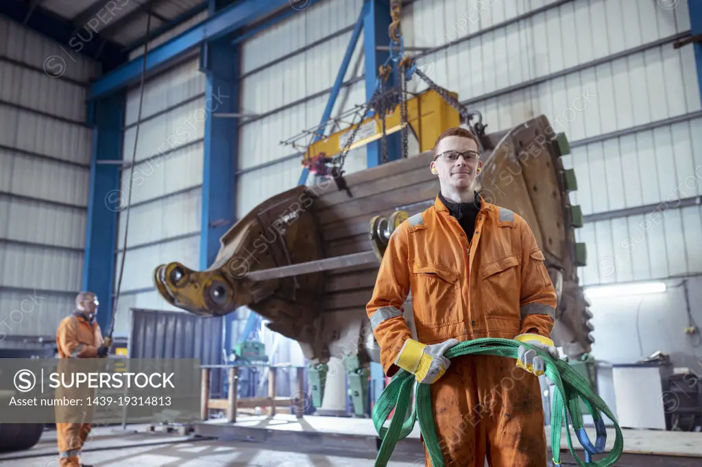 Male apprentice engineer in front of digger bucket in engineering factory