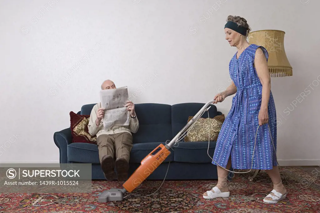 Man reading newspaper as woman vacuums