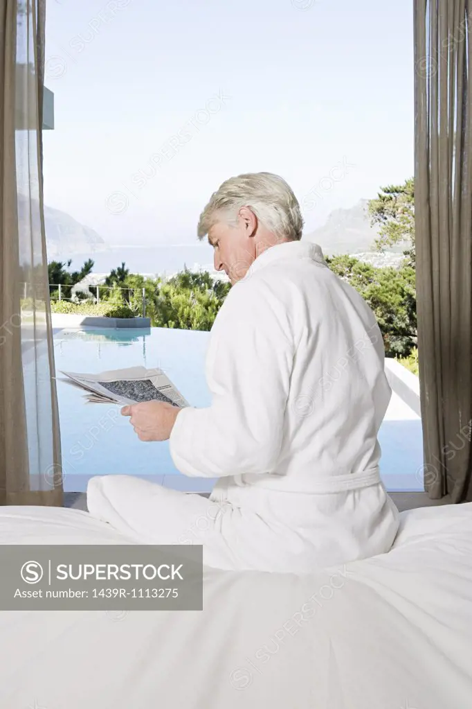 Middle aged man in robe sitting on bed reading newspaper with view out to swimming pool
