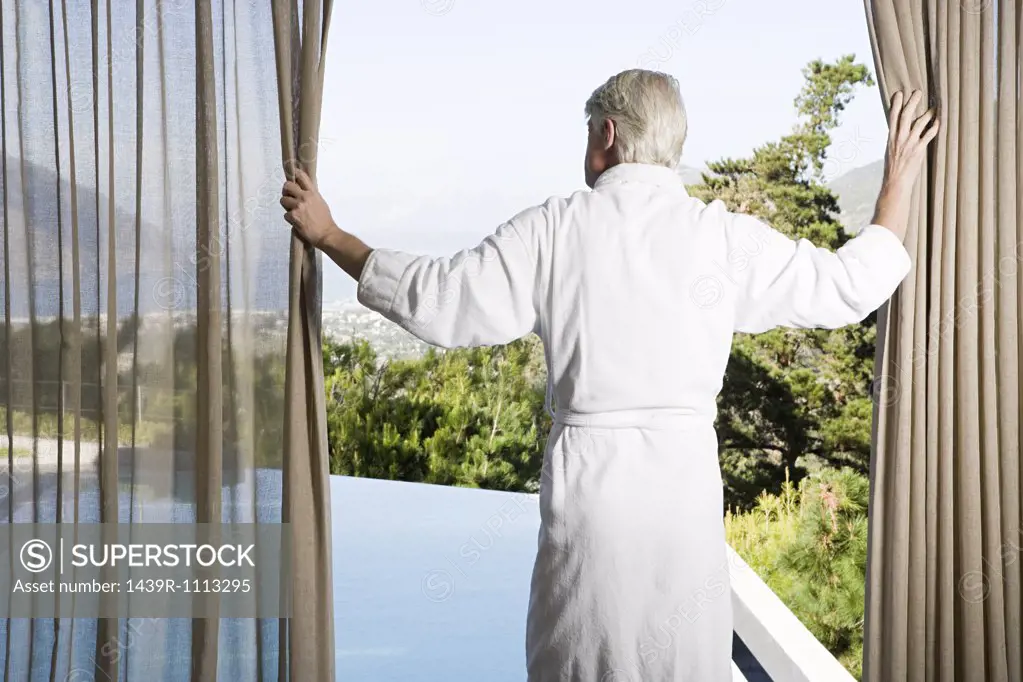 Middle aged man wearing bathrobe pulling back curtains in front of swimming pool