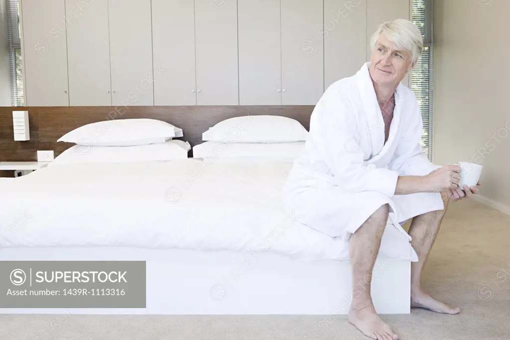 Middle aged man in bathrobe sitting on bed with cup of coffee