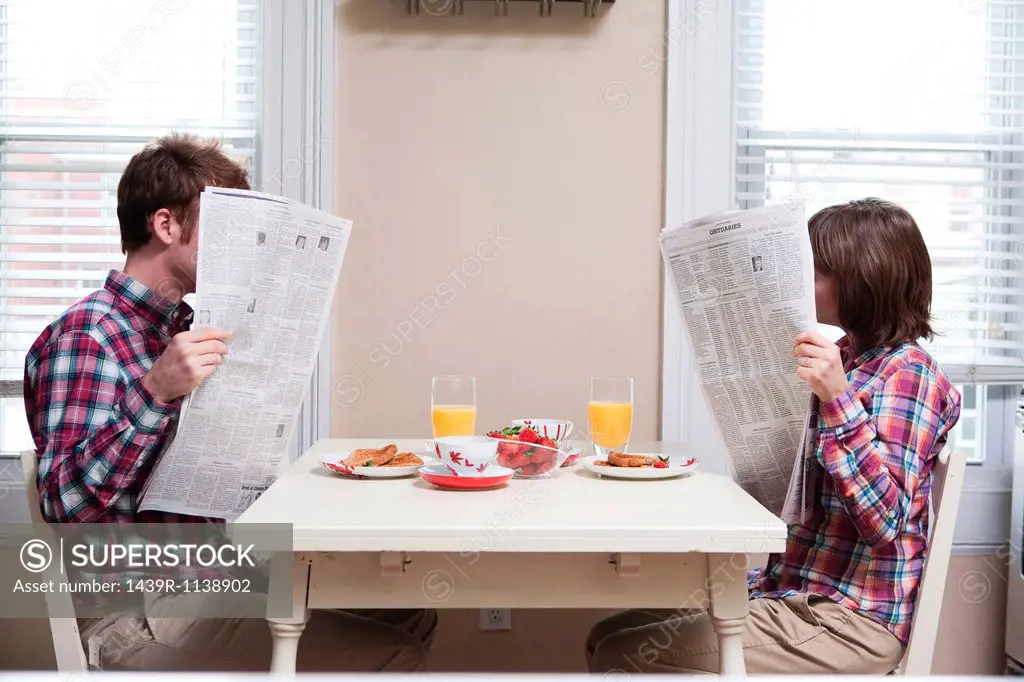 Young couple reading newspapers at kitchen table