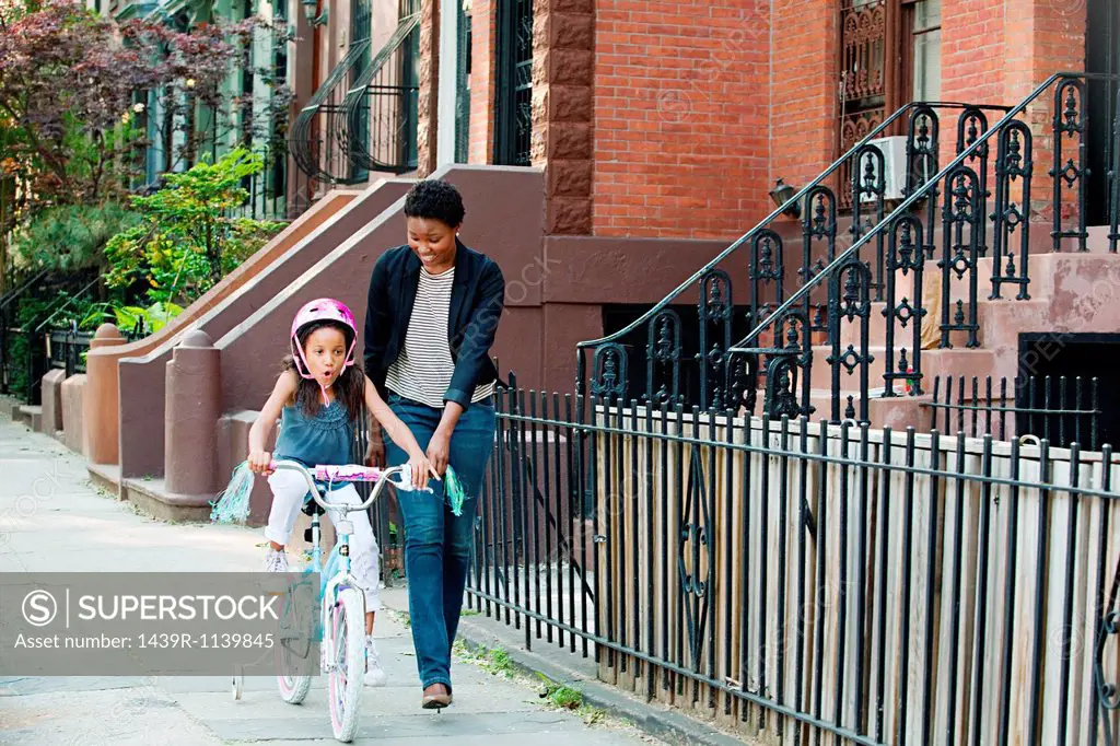 Daughter learning to ride bicycle along sidewalk with mother