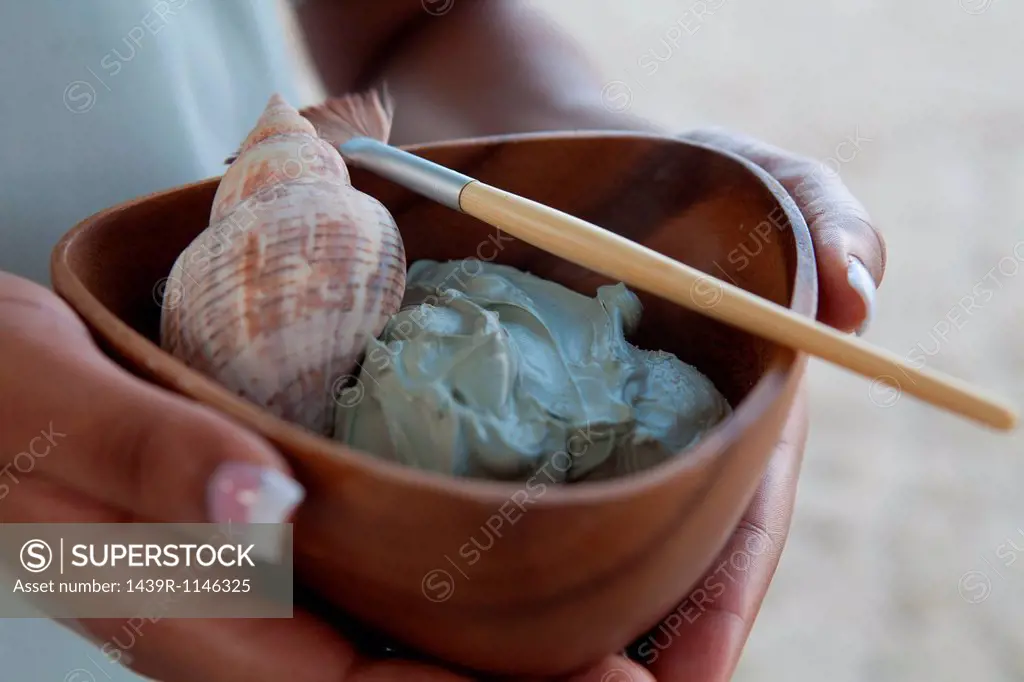 Person holding bowl of mud face mask and shell at spa