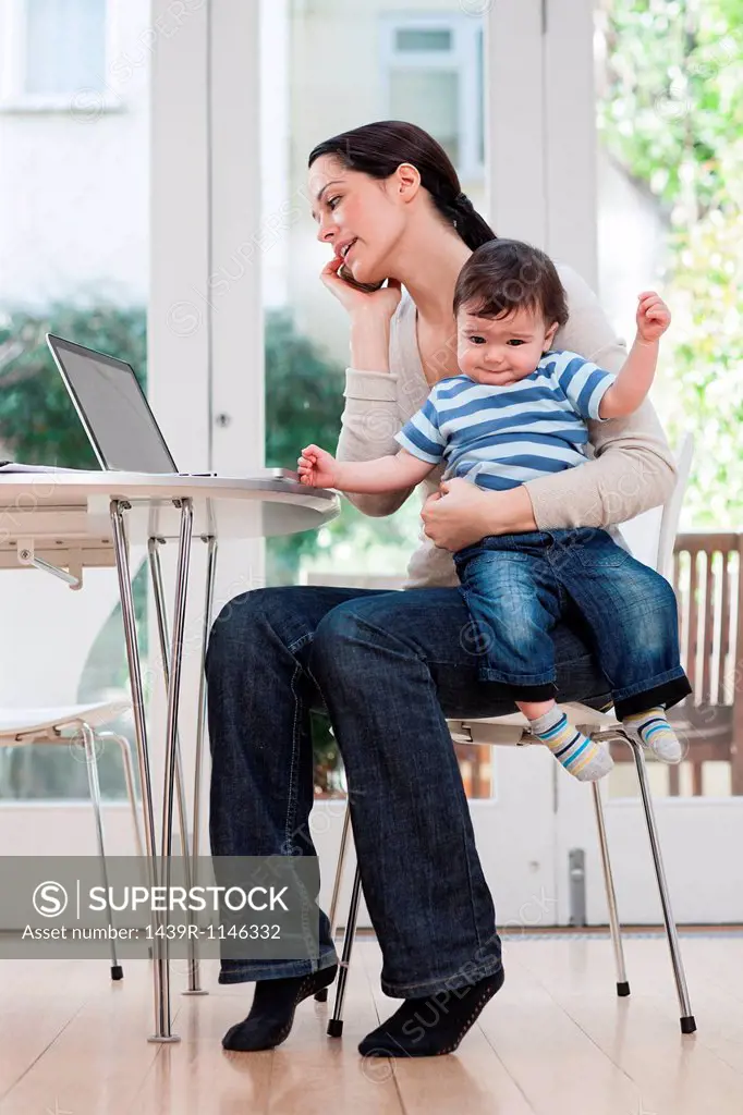 Mother holding baby boy, using cellphone and laptop