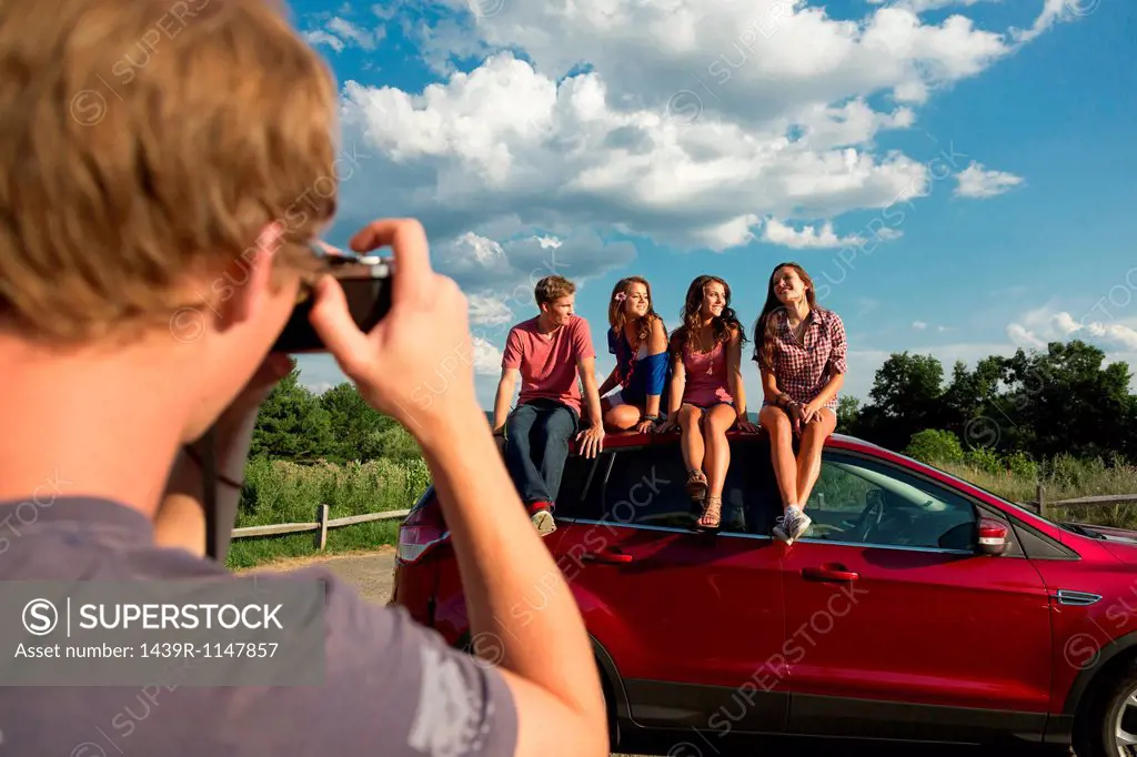 Friends sitting on car roof, young man taking photograph