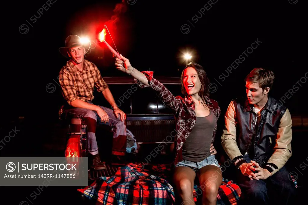 Three friends sitting on tailgate of car at night, girl holding sparkler