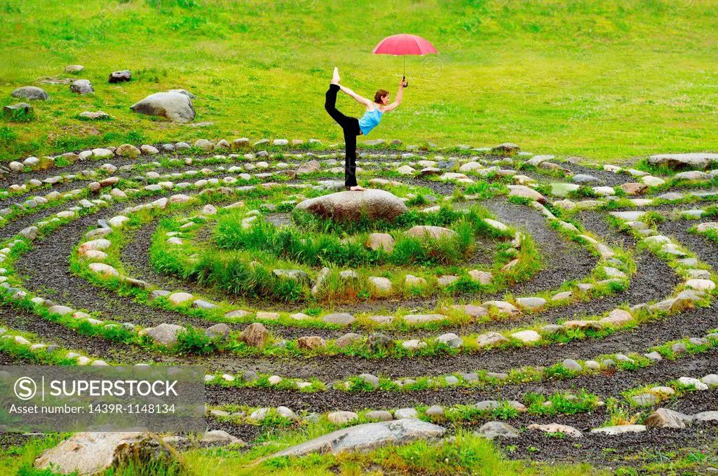 Woman holding an umbrella whilst in yoga pose in centre of stone maze