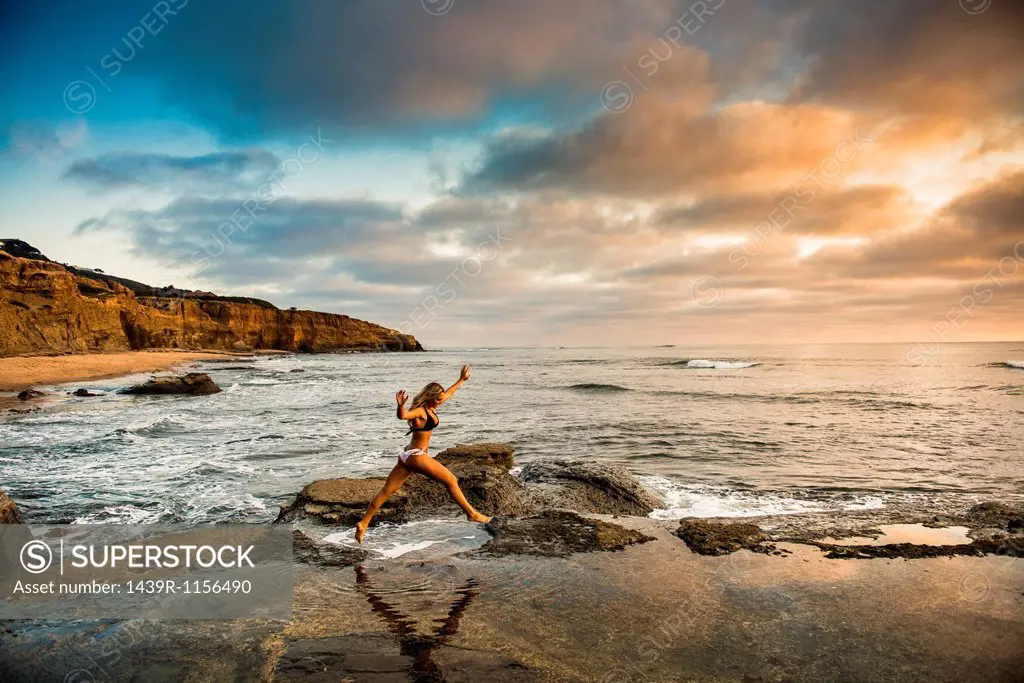 Young woman in bikini leaping from rocks on beach
