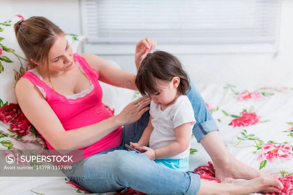 Pregnant woman and toddler daughter on bed looking at smartphone