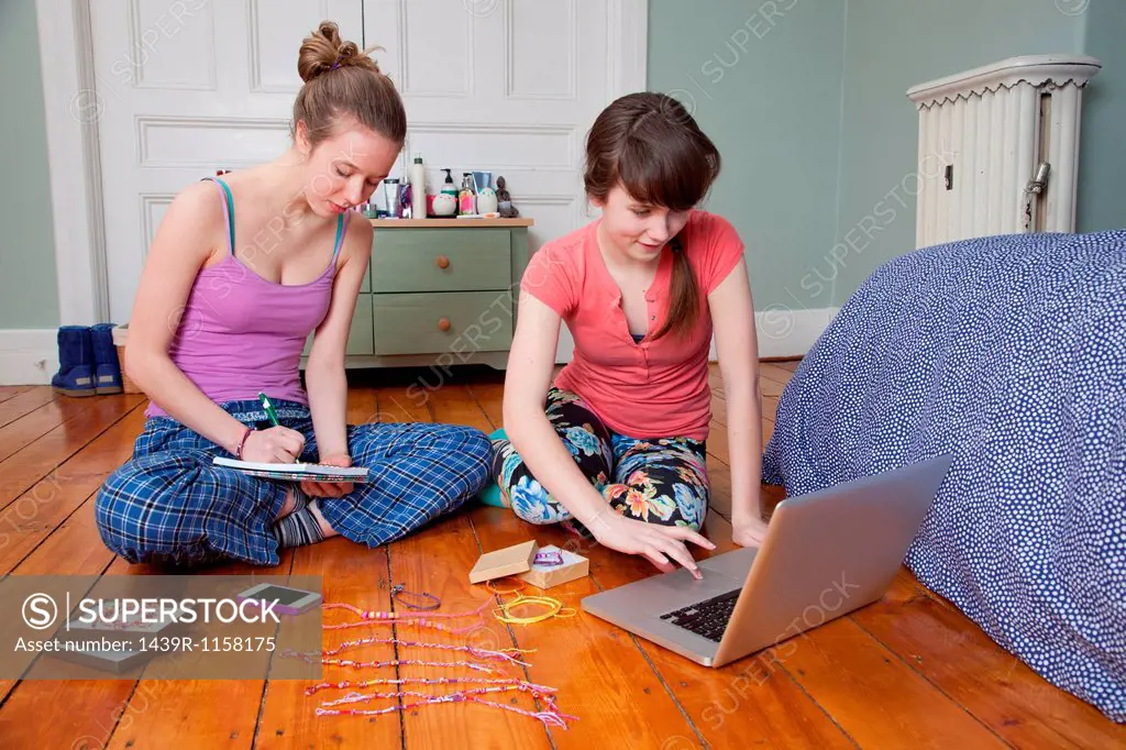 Girls sitting on bedroom floor with laptop, making friendship bracelets