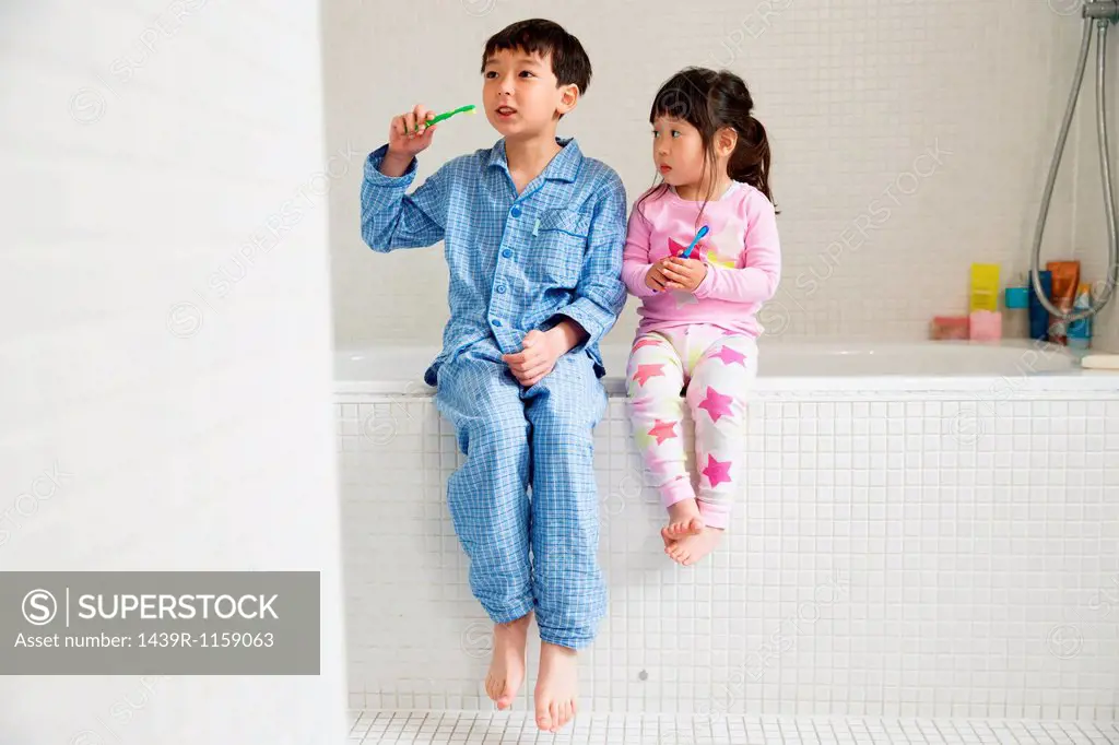 Brother and sister sitting on edge of bath with toothbrushes