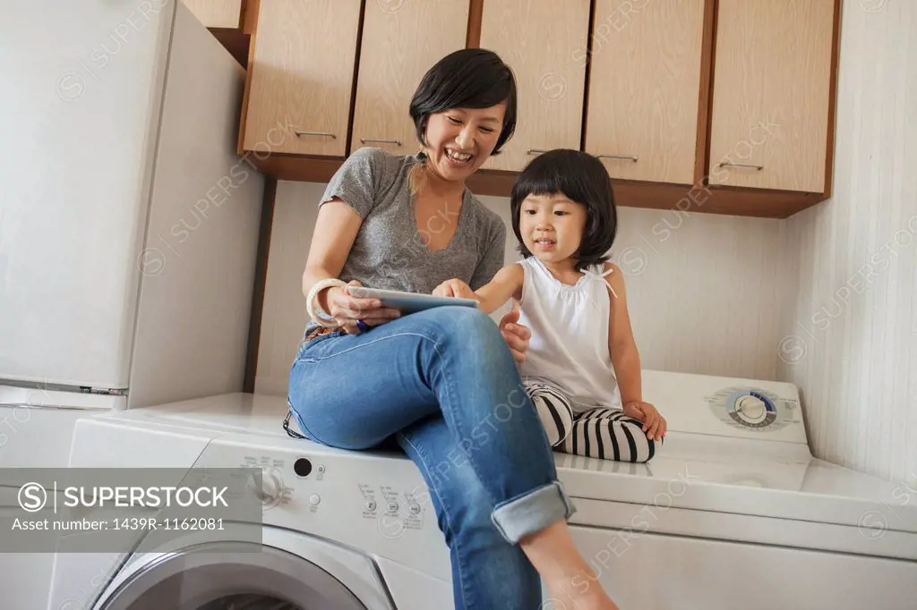 Mother and daughter sitting on washing machine using tablet