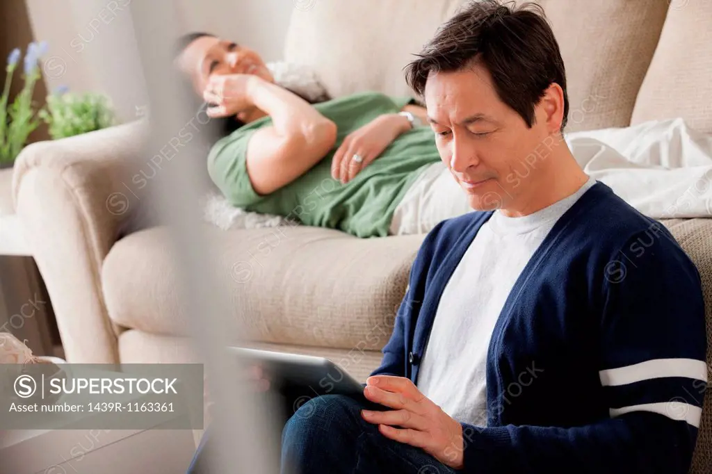 Mature man using digital tablet, woman lying on sofa