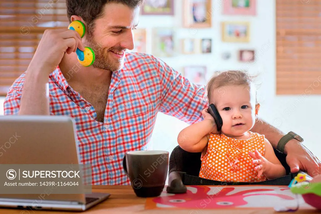 Man and baby sitting at kitchen counter playing with smartphone and toy phone