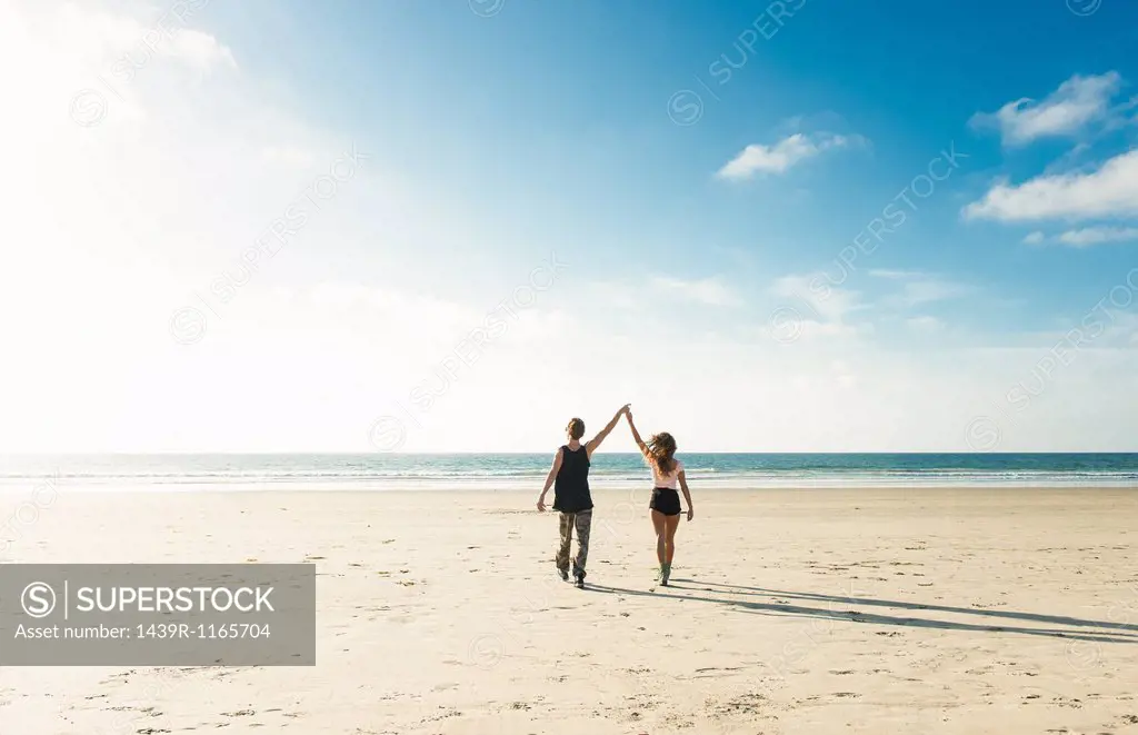 Young couple walking on San Diego beach, holding hands with arms raised
