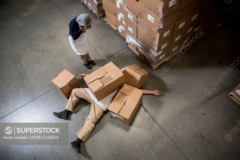 Woman looking at man lying on floor covered by cardboard boxes in warehouse