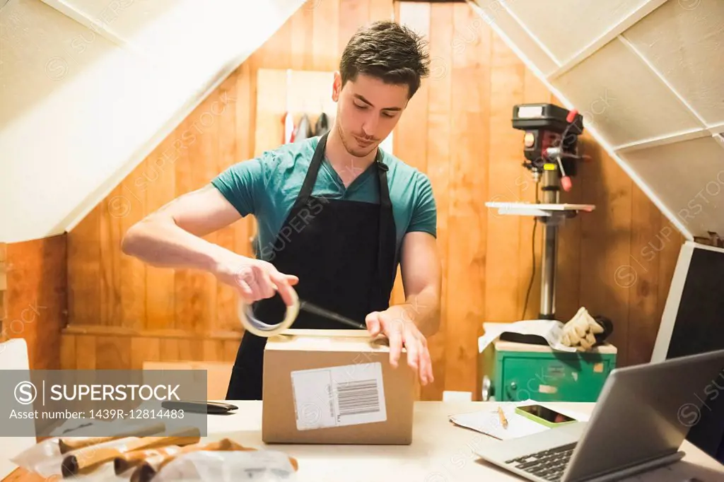 Young man using sticking tape to prepare package for delivery