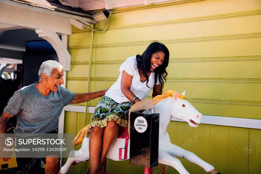 Senior woman on horse funfair ride, senior man standing beside her, laughing, Long Beach, California, USA
