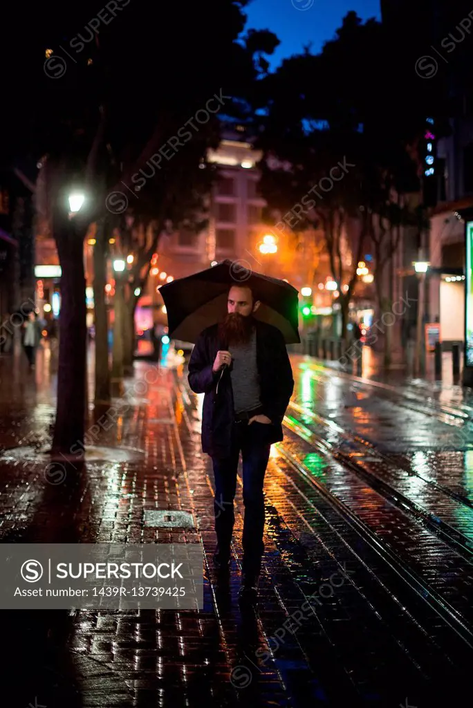 Mid adult man walking in city at night, using umbrella, Downtown, San Francisco, California, USA