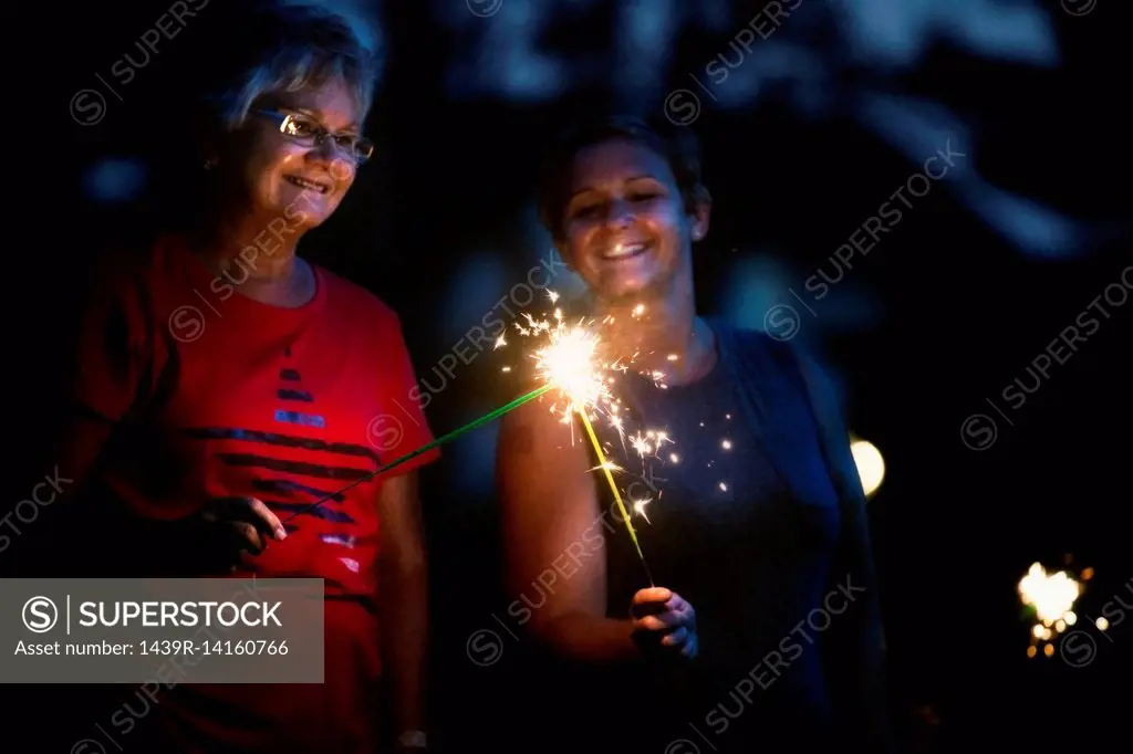 Senior and mature woman igniting sparklers together at night on independence day, USA