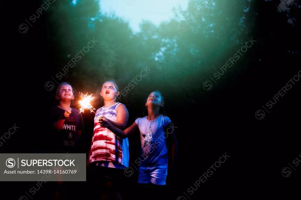 Three girls holding sparklers looking up from garden at night on independence day, USA