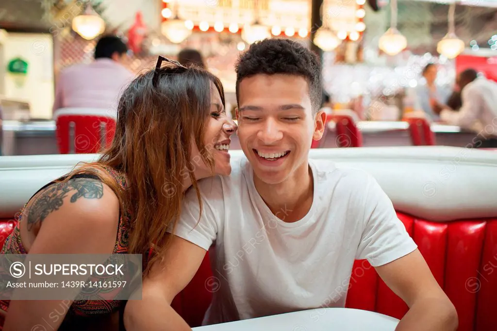 Young couple sitting in diner, young woman whispering in man's ear, laughing