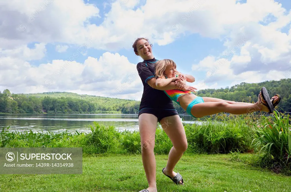 Girl swinging young girl around on grass, beside lake