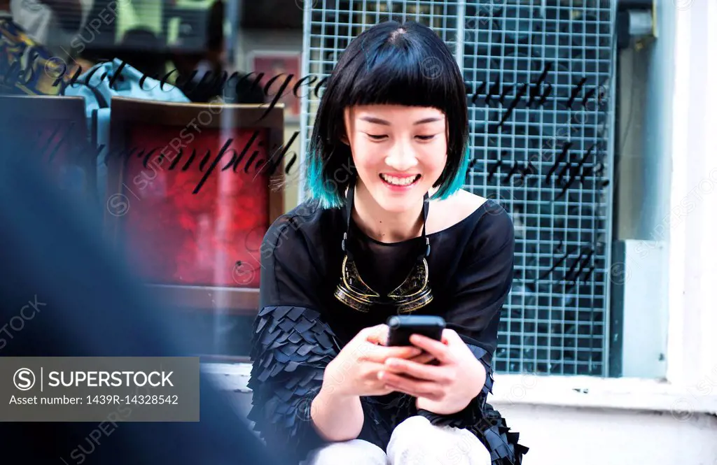 Young stylish woman sitting outside shop looking at smartphone