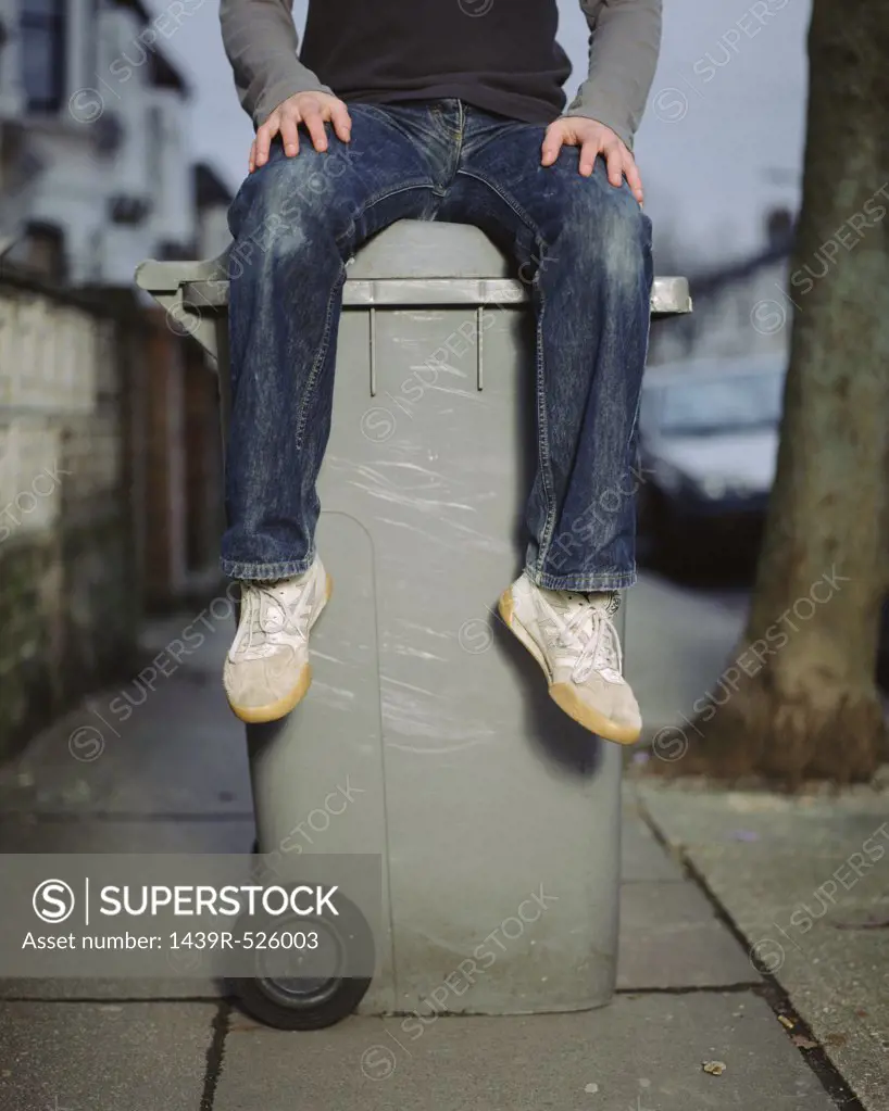 A young man sitting on a wheelie bin