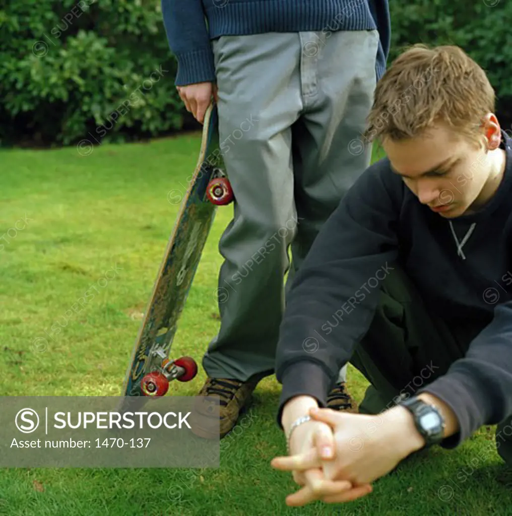Teenage boy crouching on the grass with another teenage boy standing beside him holding a skateboard