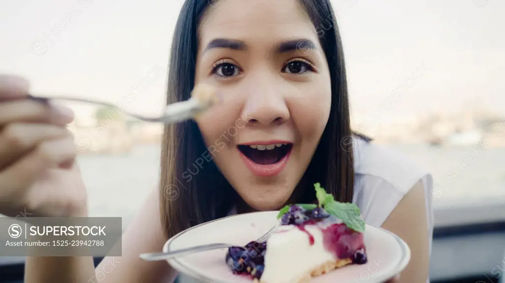Blogger Asian friendly woman influencer eating cake at cafe. Young lady happy relax fun using technology mobile phone record vlog video upload in social media holiday travel near river in city.