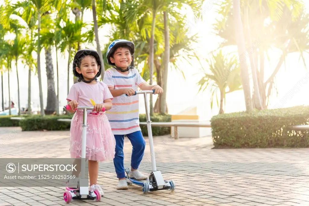 Child riding scooter. Happy Asian little kid boy and girl wear safe helmet play kick board on road in park outdoors on summer day, Active children games outside, Kids sport healthy lifestyle concept