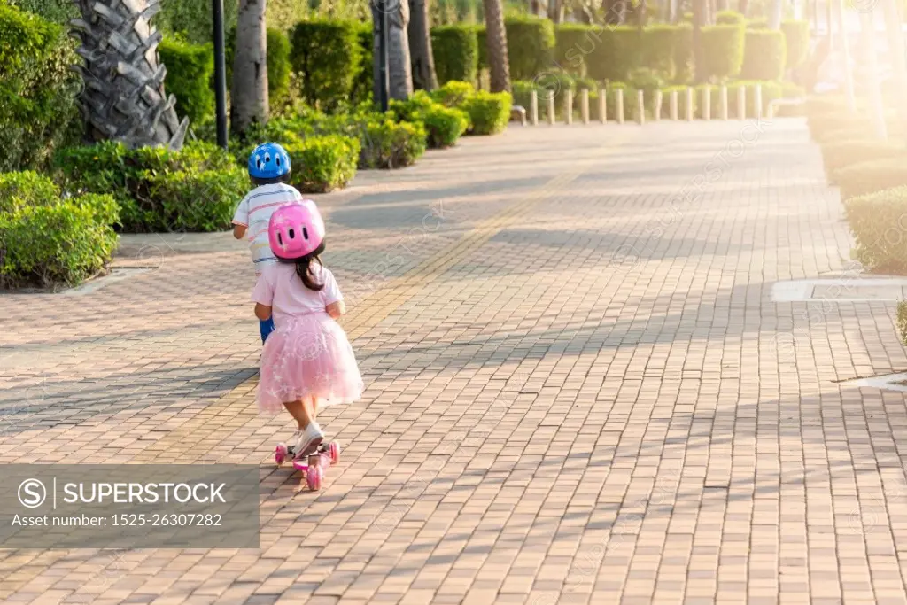 Child riding scooter. Back Asian little kid boy and girl wear safe helmet play kick board on road in park outdoors on summer day, Active children games outside, Kids sport healthy lifestyle concept
