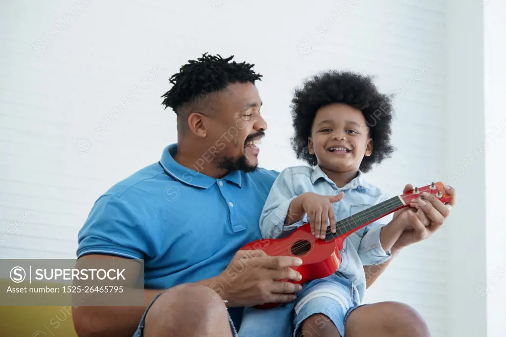 African American father and little son singing and playing ukulele together. Dad teaching his boy play a small red ukulele on sofa at home