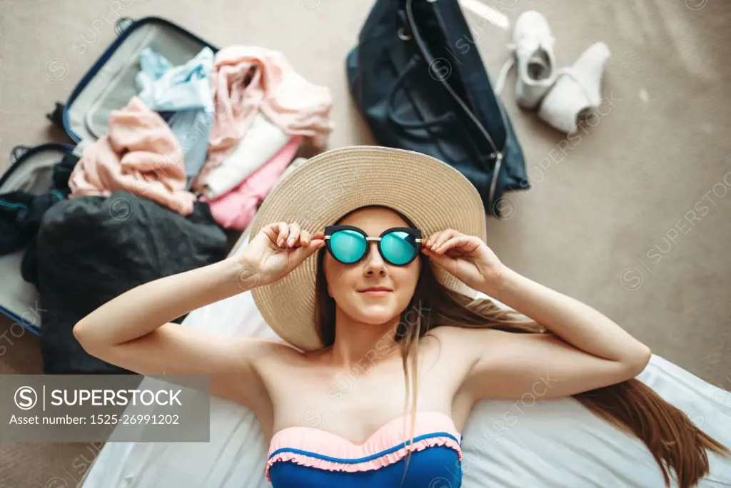 Young woman lies in swimsuit and sunglasses, top view, opened suitcase with clothes on background. Fees on journey concept. Preparation for summer vacation. Woman lies in swimsuit and sunglasses, top view