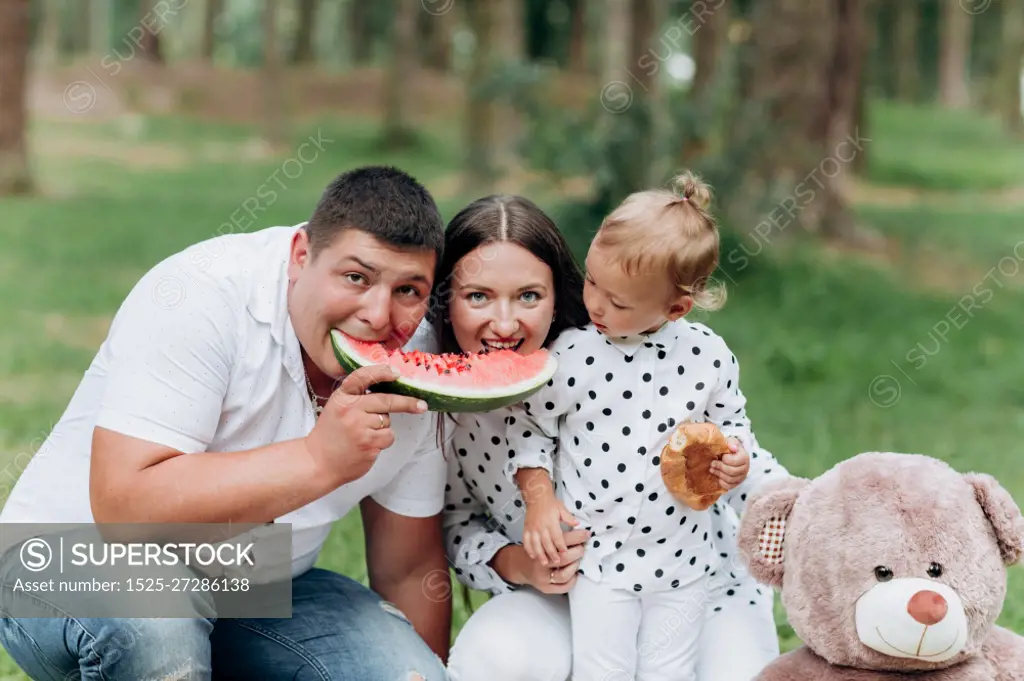 holiday outdoor portrait of happy families eating watermelon at the picnic. mom, dad and daughter with her teddy having fun in the park together. selective focus.. holiday outdoor portrait of happy families eating watermelon at the picnic. mom, dad and daughter with her teddy having fun in the park together. selective focus