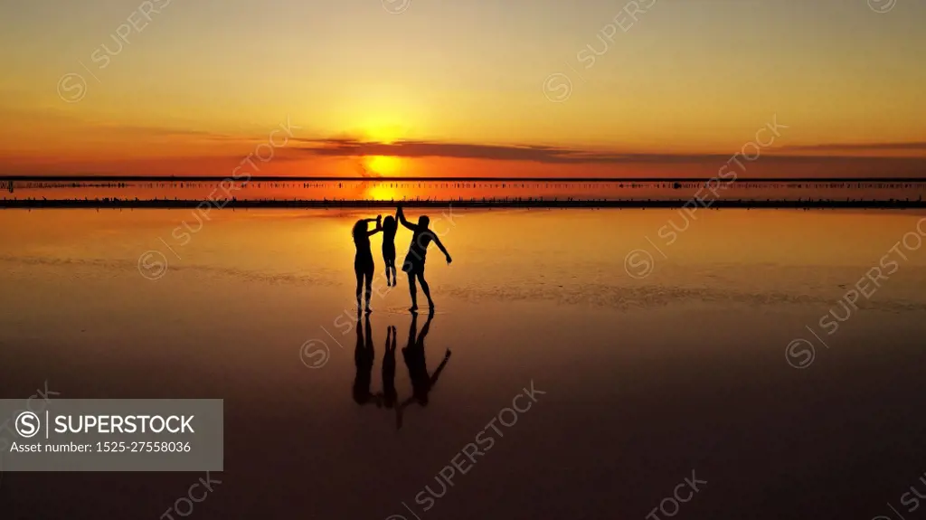 Silhouettes of three, dad, mom and little daughter walking through the shallows of a lake against the background of a red-orange sunset.. Silhouettes of three, dad, mom and little daughter walking through the shallows of a lake against the background of a red-orange sunset