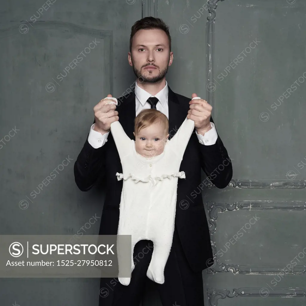 Young father with beautiful little baby in his arms. Fashion portrait of man with pretty daughter. Stylish dad in evening black suit and small girl in white sliders. Studio portrait.