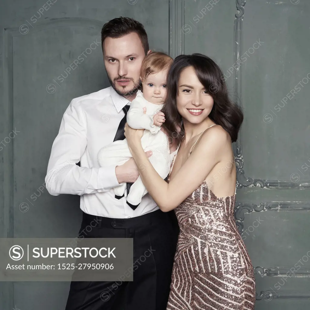 Young family with beautiful little baby in thair arms. Fashion portrait of two parents with pretty daughter. Stylish mom and dad in evening dress and small girl in white sliders. Studio portrait.