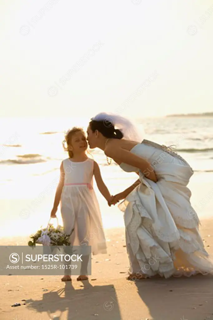 Caucasian mid-adult bride kneeling to give flower girl a kiss on the cheek while holding hands barefoot on beach.