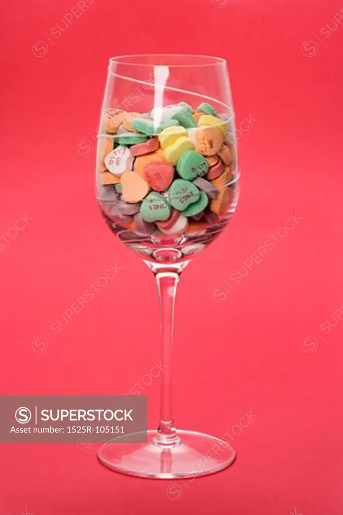 Wine glass full of colorful candy hearts with sayings on them on red background.