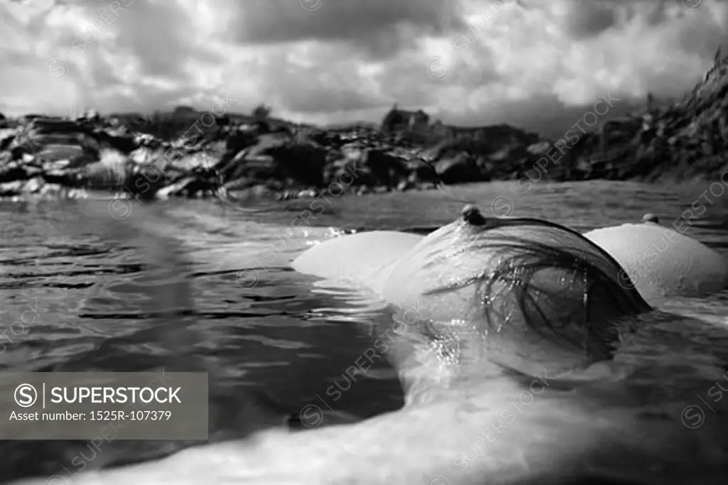 Breast of young Asian nude woman floating on back in water on rocky coast in Hawaii.