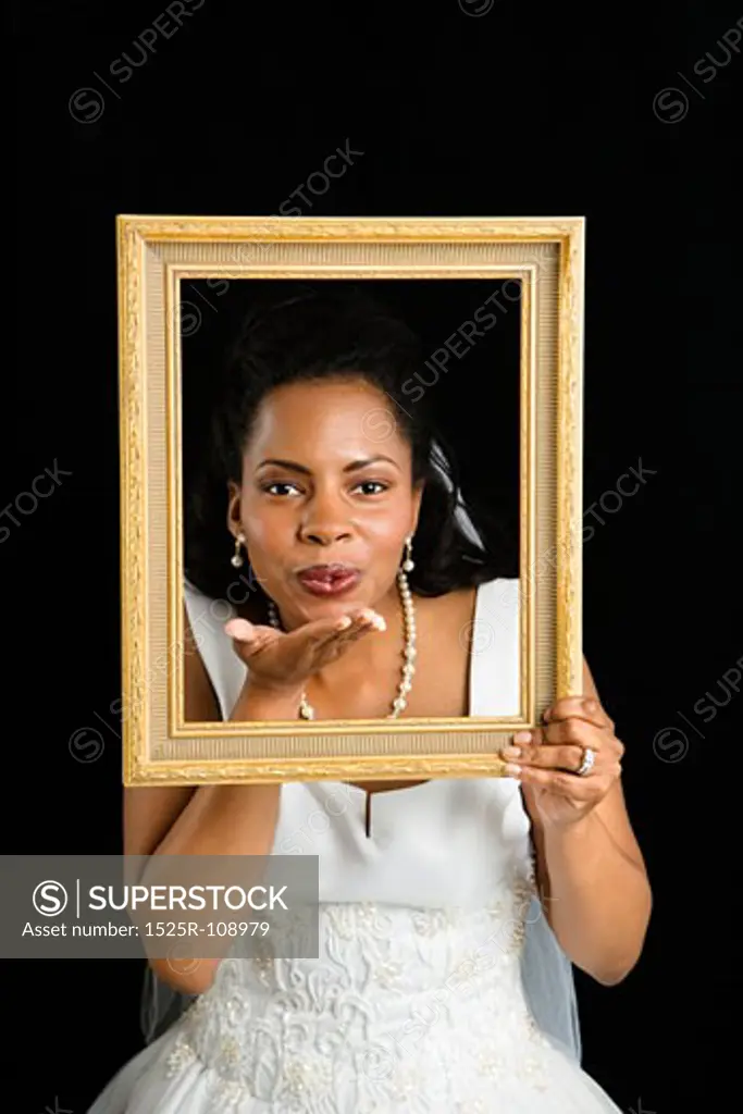 Mid-adult African-American bride holding frame around her face and blowing a kiss.