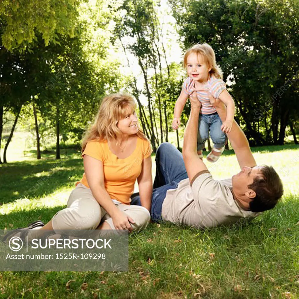 Caucasian mid adult parents holding toddler daughter in the air playing at grassy park.