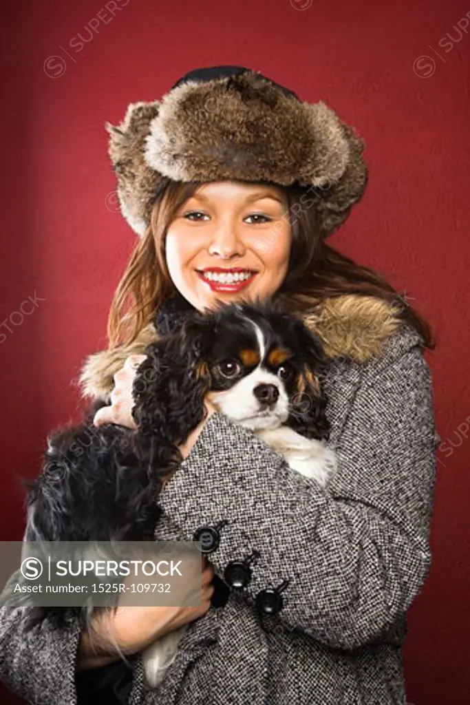 Young adult Caucasian woman wearing fur hat holding King Charles Spaniel in arms and smiling at viewer.