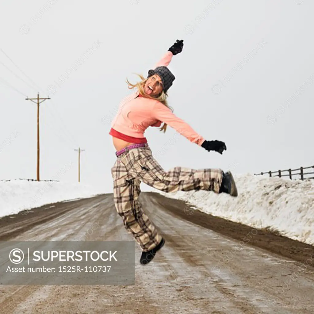 Young woman in winter clothes standing on muddy dirt road jumping and smiling.