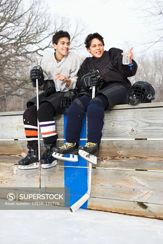 Two boys in ice hockey uniforms sitting on ice rink sidelines pointing and looking.